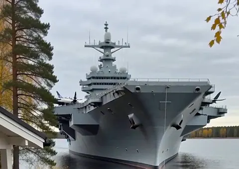 Harmaa lentotukialus järvellä aivan mökkirannan edustalla, etualalla männyt ja tyyni vesi.