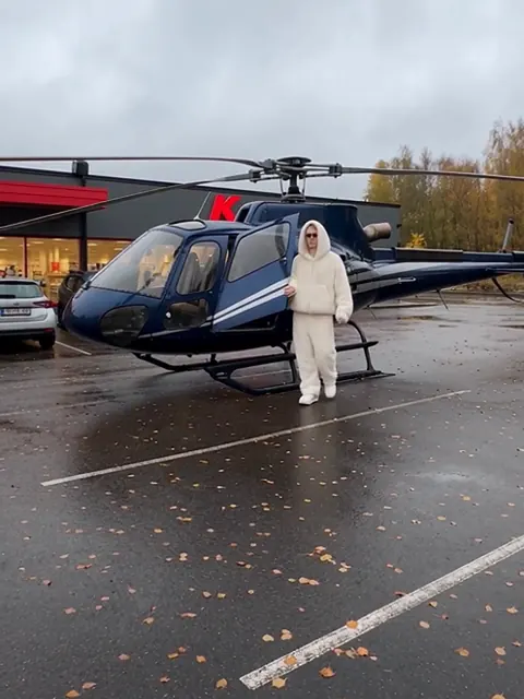 Tummansininen helikopteri pysäköitynä marketin märälle parkkipaikalle henkilöautojen viereen, valkoisiin pukeutunut hahmo kävelee ohi.